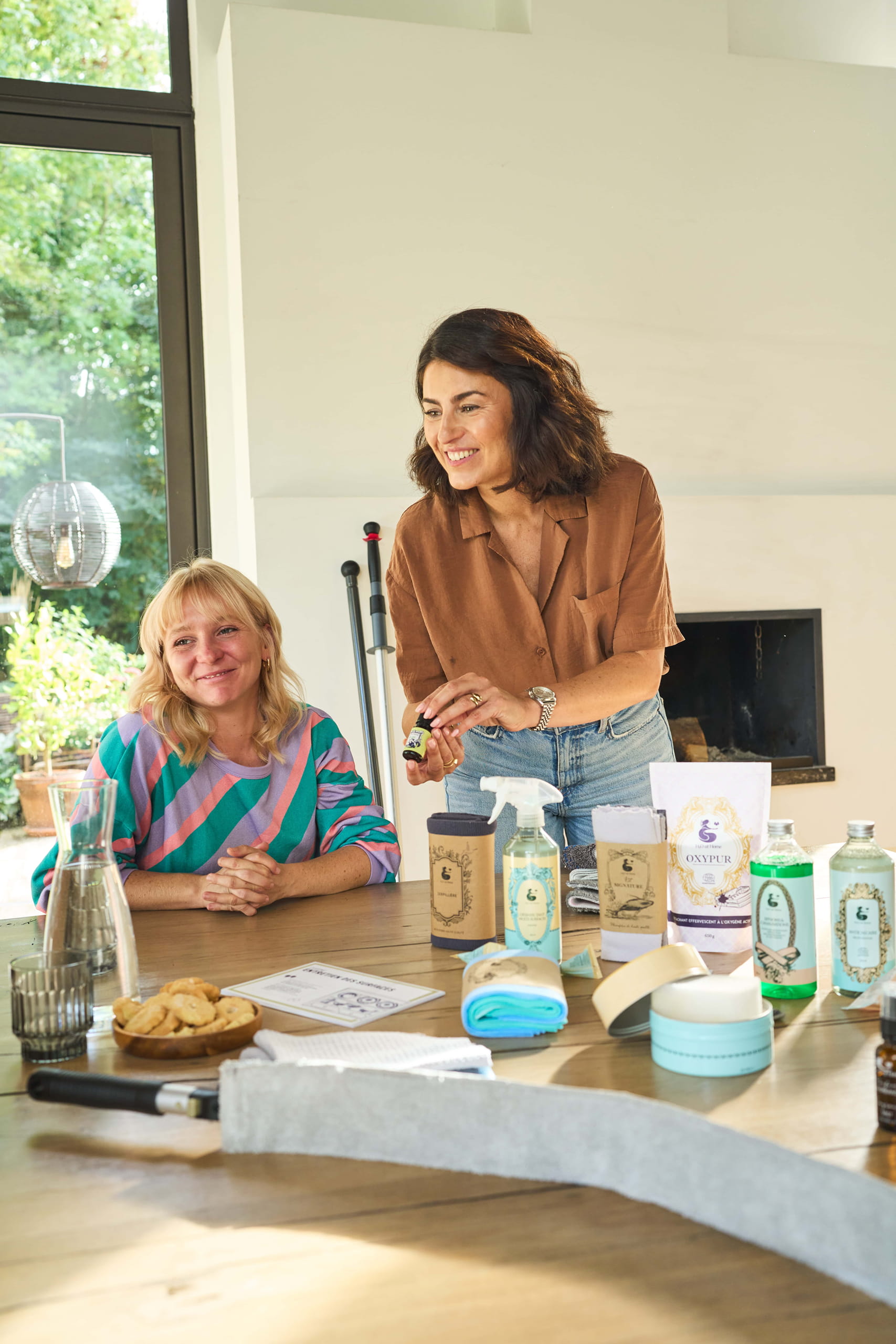 Photo d'un atelier H2O at Home à domicile avec une conseillère montrant un produit Synergie avec de nombreux autres produits sur la table