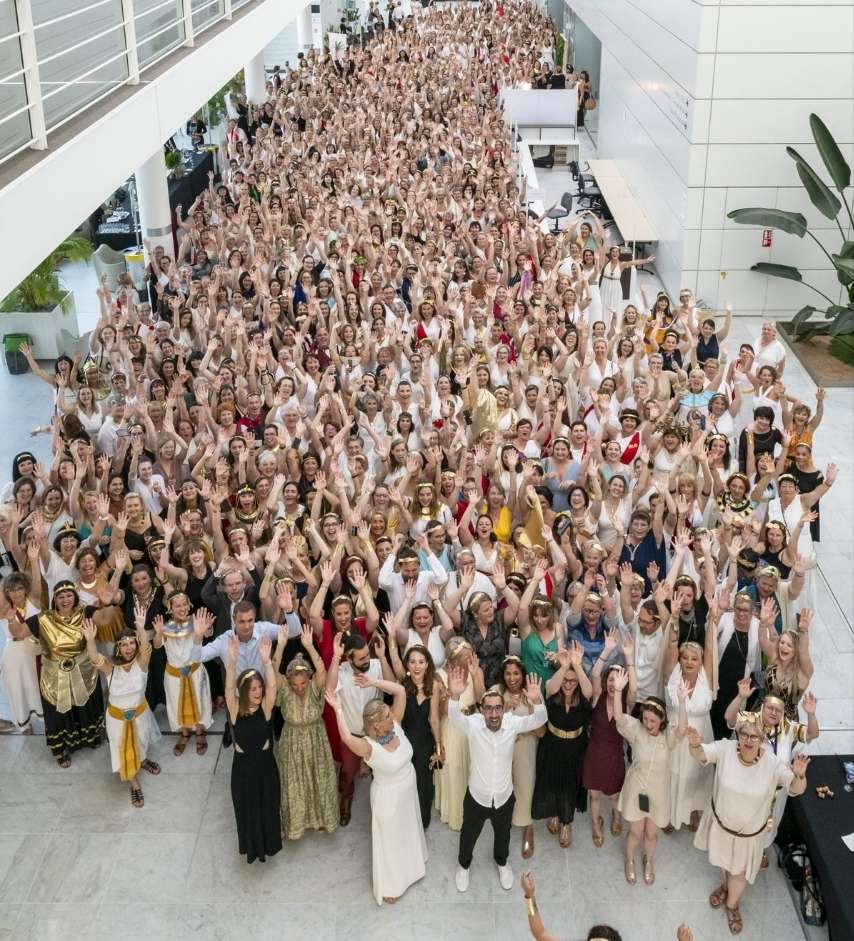 Grande assemblée de personnes en tenue antique, majoritairement blanche et dorée, rassemblées dans un hall moderne et levant les bras dans une ambiance festive