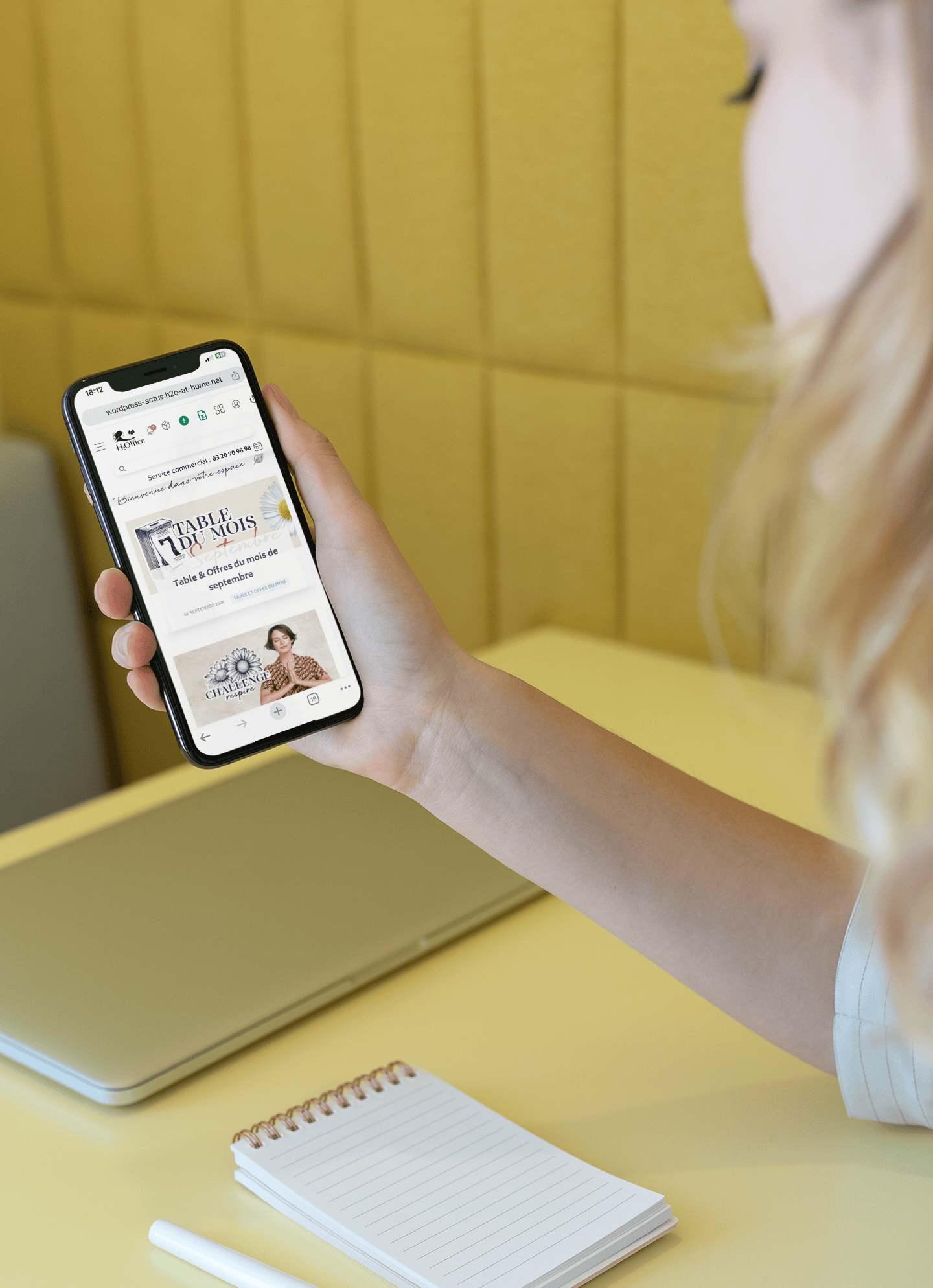 Femme blonde tenant un iPhone affichant une page web avec des offres promotionnelles. Elle est assise à un bureau avec un ordinateur portable fermé, un carnet et un stylo posés devant elle, dans un cadre moderne et lumineux